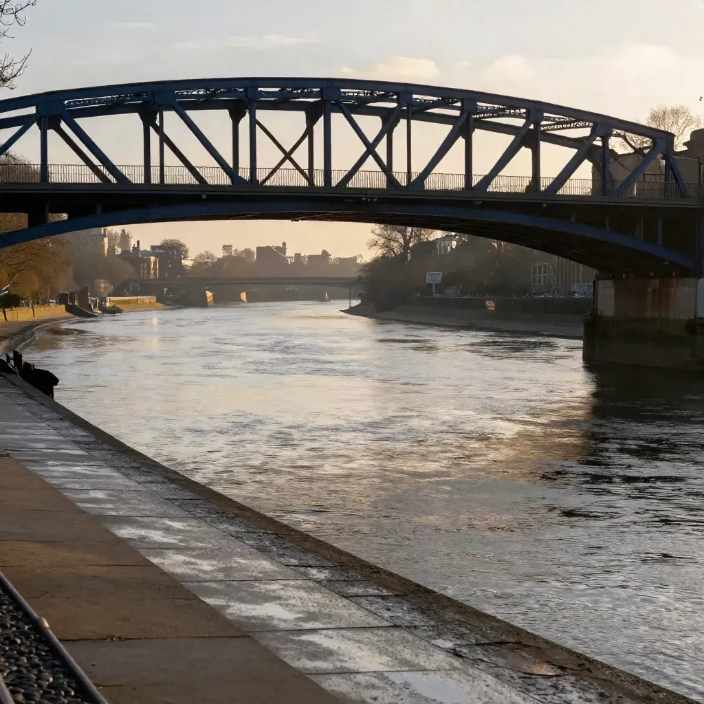 Hammersmith Bridge over the Thames in warm mist at sunset, with calm water and wet riverbank.